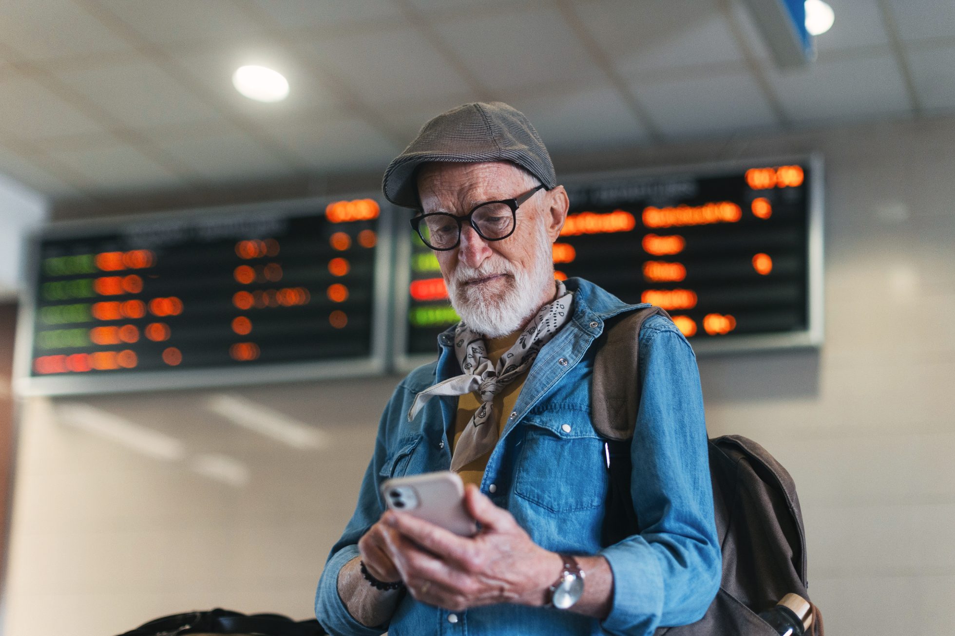 A stylish elderly man at the airport under the departure board, checking his reservation on a smartphone.